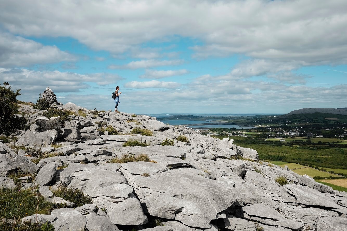 PNCA student studying abroad in Ireland overlooking green land and water on a mountain of rocks