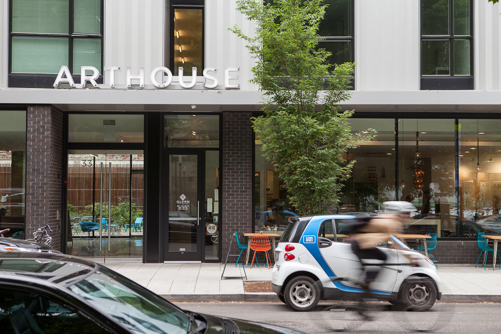 The facade of a modern building with "ARTHOUSE" sign on top, a glass front with visible indoor seating area, a small electric car parked by the curb, and a cyclist in motion passing by on a city street.