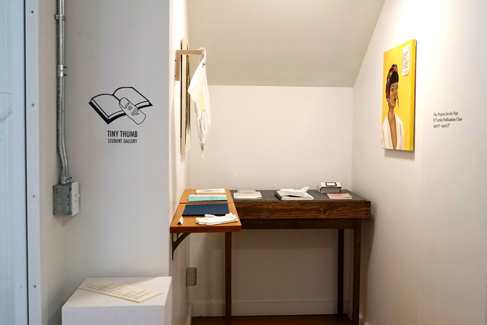 A photo of a student gallery titled Tiny Tumb, featuring a painting of a woman with a yellow background and a desk with papers