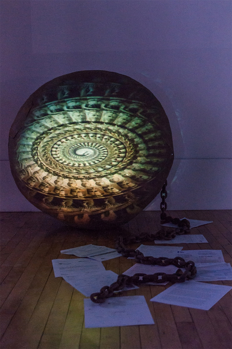Exhibition featuring paper on the floor and a chain leading up to a large circle with green and yellow patterns