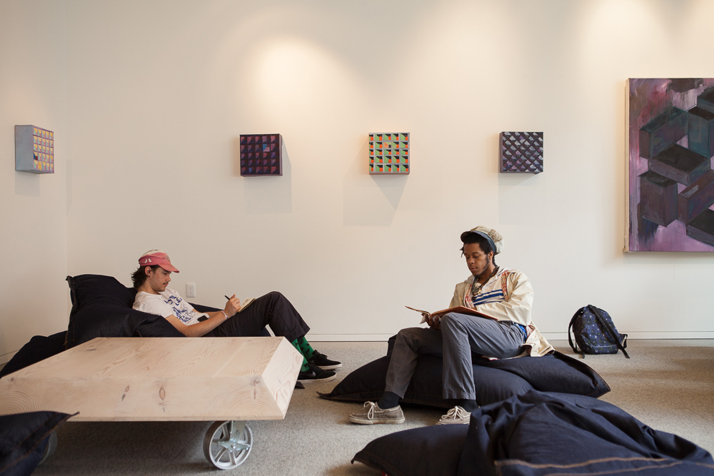 Two people seated on black beanbags in a serene room with abstract art on the walls, one using a mobile device and the other reading a book.