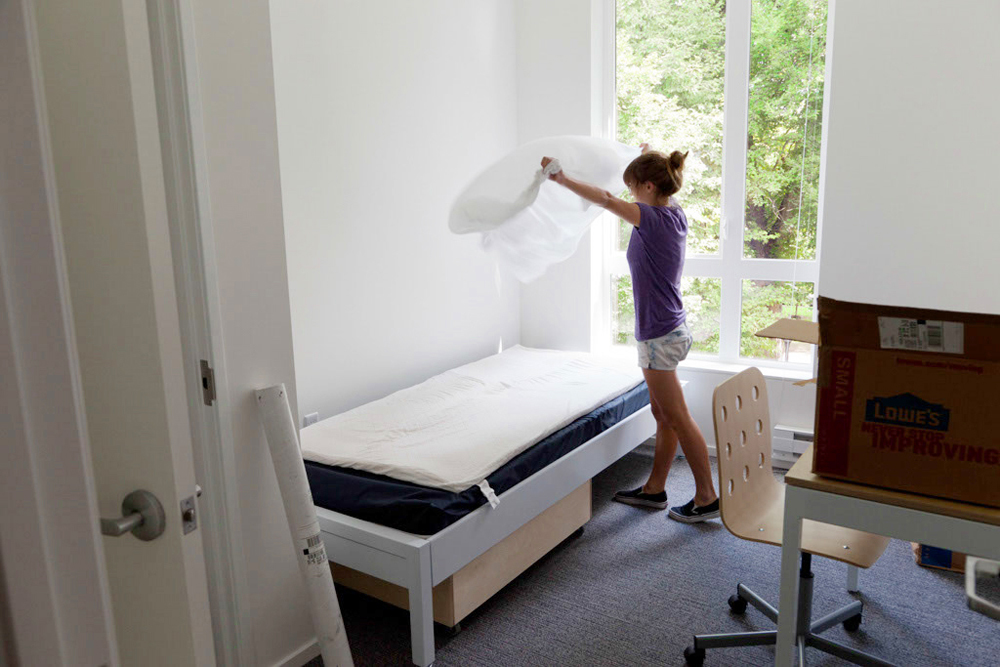 A person is making a bed by placing a white sheet on it in a bright room with a window, desk, chair, and an open door, with a moving box on the floor.