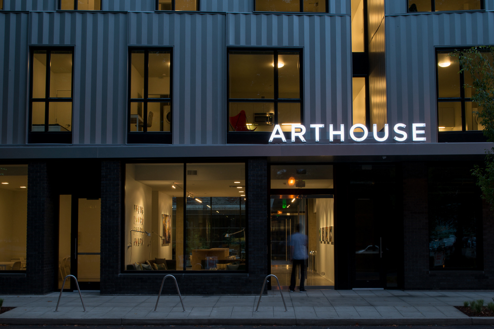 Twilight view of a modern building named ARTHOUSE, showing illuminated signage above the main entrance, with a blurred figure walking inside, and a warmly lit interior visible through large windows.