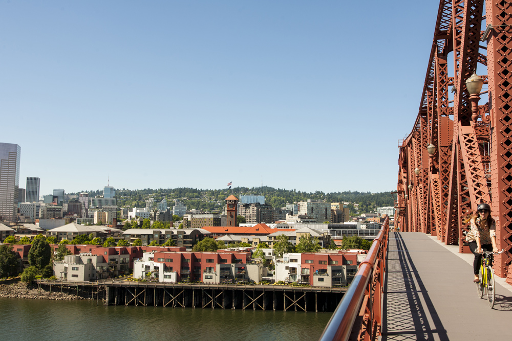 Portland's red-colored Broadway Bridge on a sunny day with downtown Portland in the distance and a bike rider on the walkway.