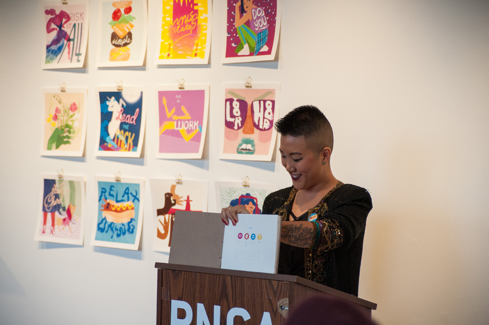 Studen Jax Ko standing behind a podium before a display of her illustrations giving her thesis presentation during Pacific Northwest College of Art (PNCA) Focus Week 2016.