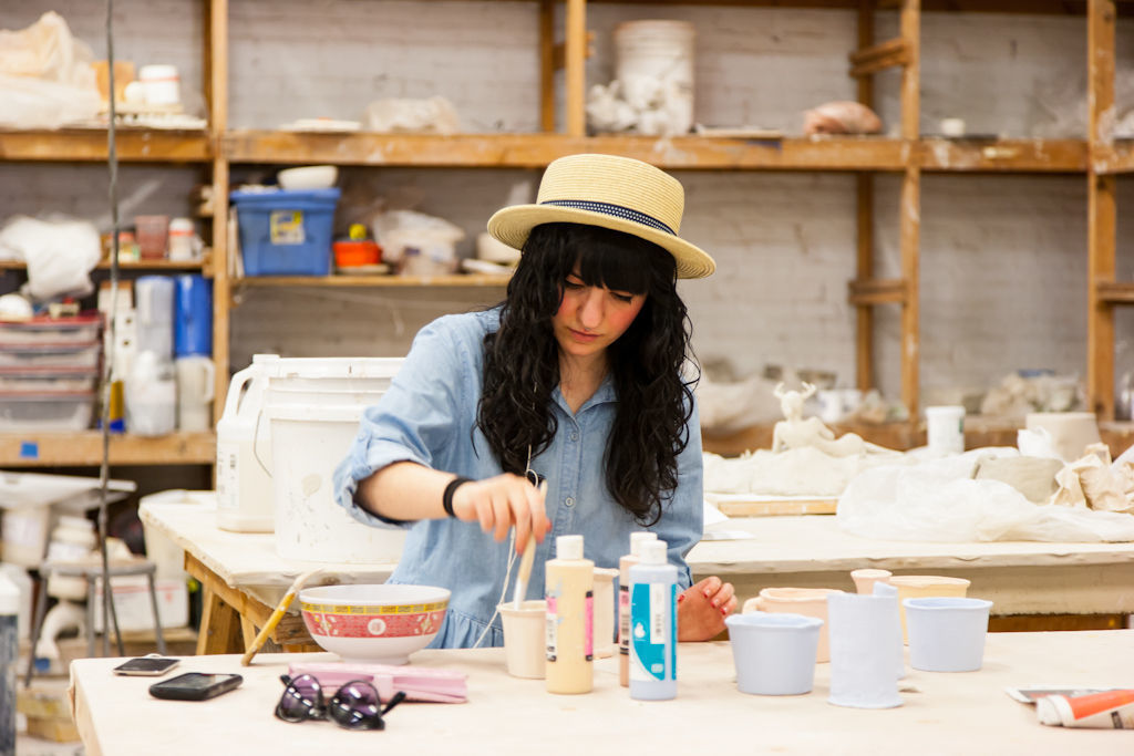 A person in a straw hat and blue denim shirt working on small white ceramic pieces in a pottery studio, with shelves of art supplies and unfinished pottery in the background.