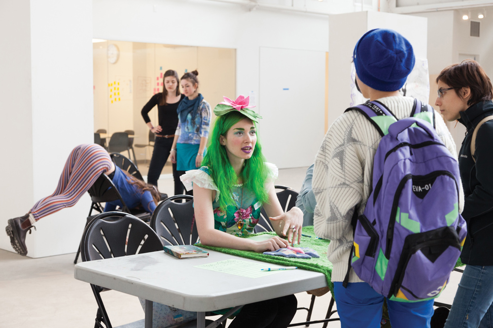 Green haired student of the Ecology Club talking with other Pacific Northwest College of Art (PNCA) students while Yoga Club students demonstrate acro yoga asanas in the background. 