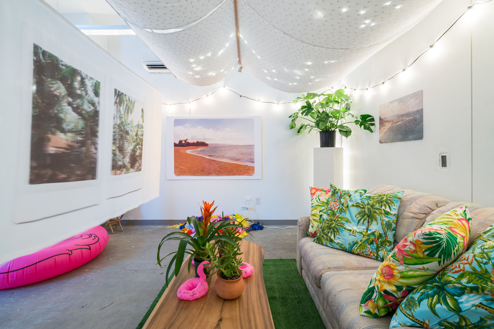 An installation of a tropical interior, featuring flamingo drink floaties, tropical plants, tropical pillows, and pictures of tropical destinations